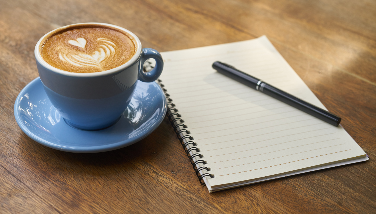 A blue cup of coffee beside a blank lined notebook and pen on a wooden desk