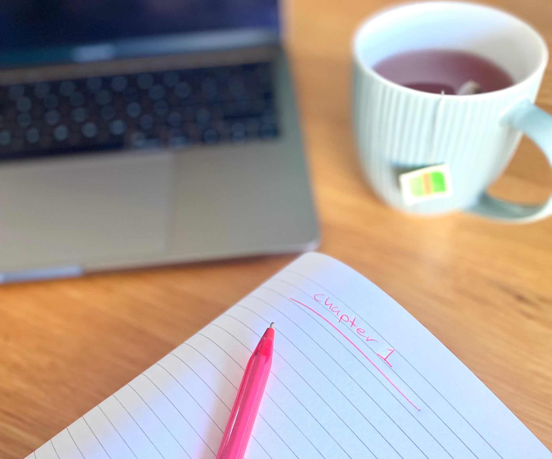 A laptop, open notebook and pink pen on a wooden table beside a cup of tea